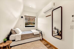 A small, neatly arranged bedroom with a single bed, a window, a mirror, and a small table.