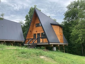 A modern, triangular wooden cabin with a balcony, surrounded by lush greenery.