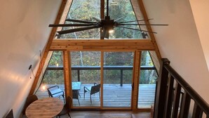 Um quarto com pé-direito alto, uma grande janela com vista para um deck, piso de madeira e um ventilador de teto.
