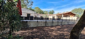 A stone wall with a metal fence, a building with a tiled roof, and trees in the background.