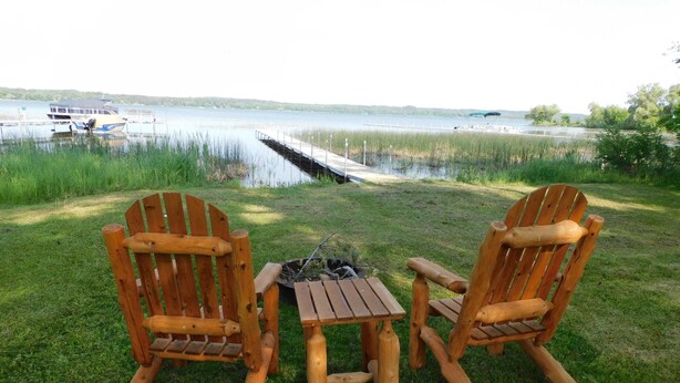 Två trästolar i Adirondack-stil och ett litet träbord står på en gräsyta vid en sjö, med en brygga och båtar i bakgrunden.