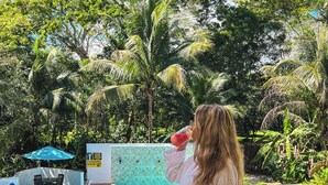 Persona con bata blanca de pie en un balcón con vista a una zona de piscina y un letrero de vestuario, rodeada de abundante vegetación.