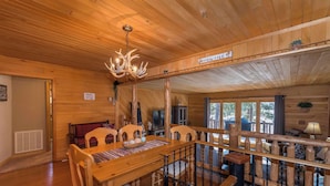 Interior de una cabaña de madera con comedor, lámpara de araña y un cartel que anuncia una experiencia llamada 'Life at the Cabin'.