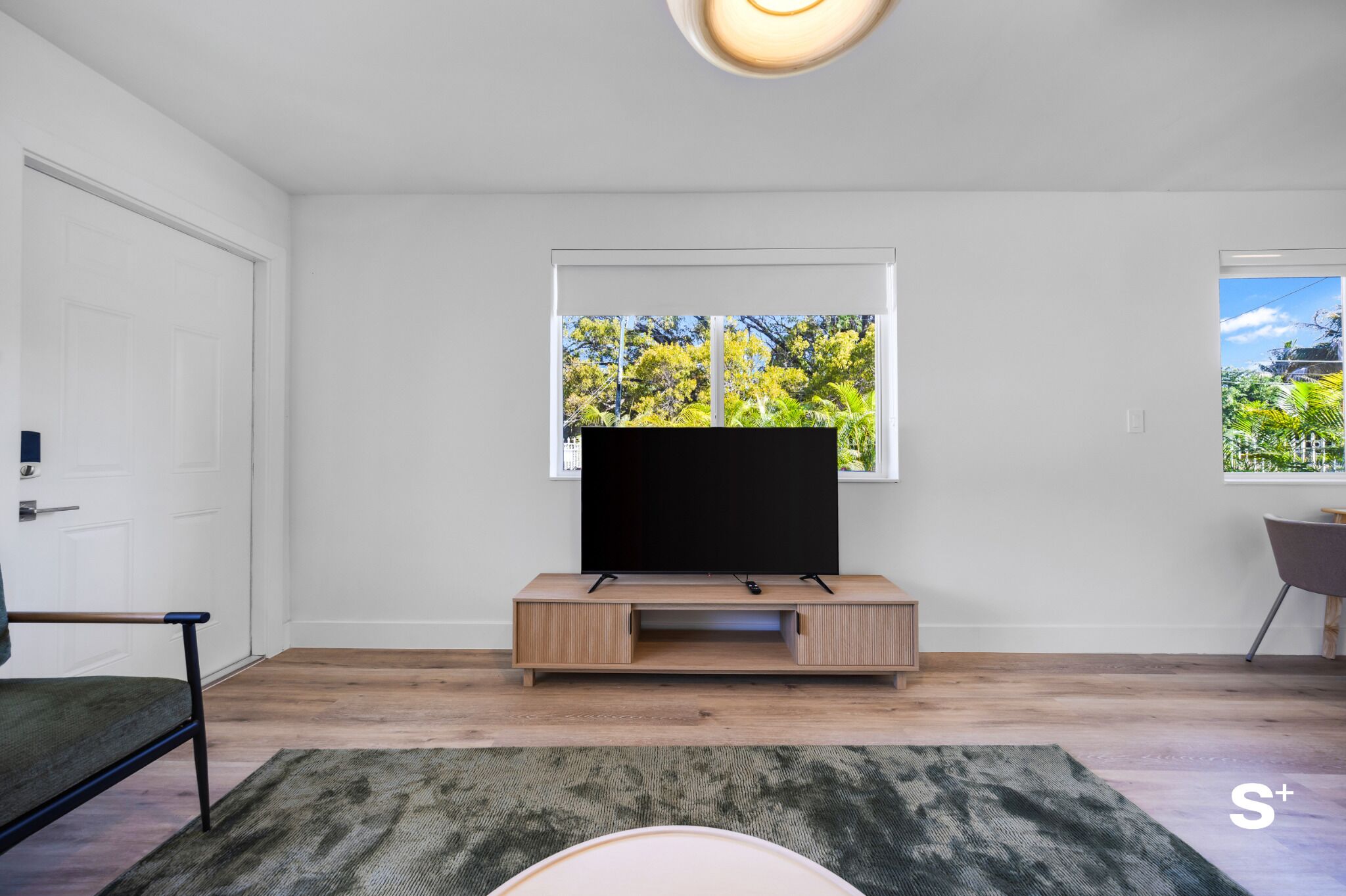 Un salon moderne avec un téléviseur à écran plat posé sur un meuble en bois, une grande fenêtre donnant sur un paysage verdoyant et un tapis vert sur le sol.