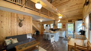 Amplio interior de cabaña con detalles de madera, una escalera, una zona de cocina y un área de descanso con sofá y mesa.
