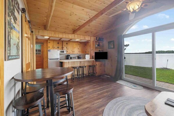 Une cabane en bois comprenant une cuisine, un coin repas et une grande porte-fenêtre coulissante donnant sur un lac.