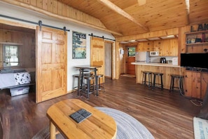Interior de una cabaña de madera con cocina, comedor y un amplio ventanal corredera que da a un dormitorio.