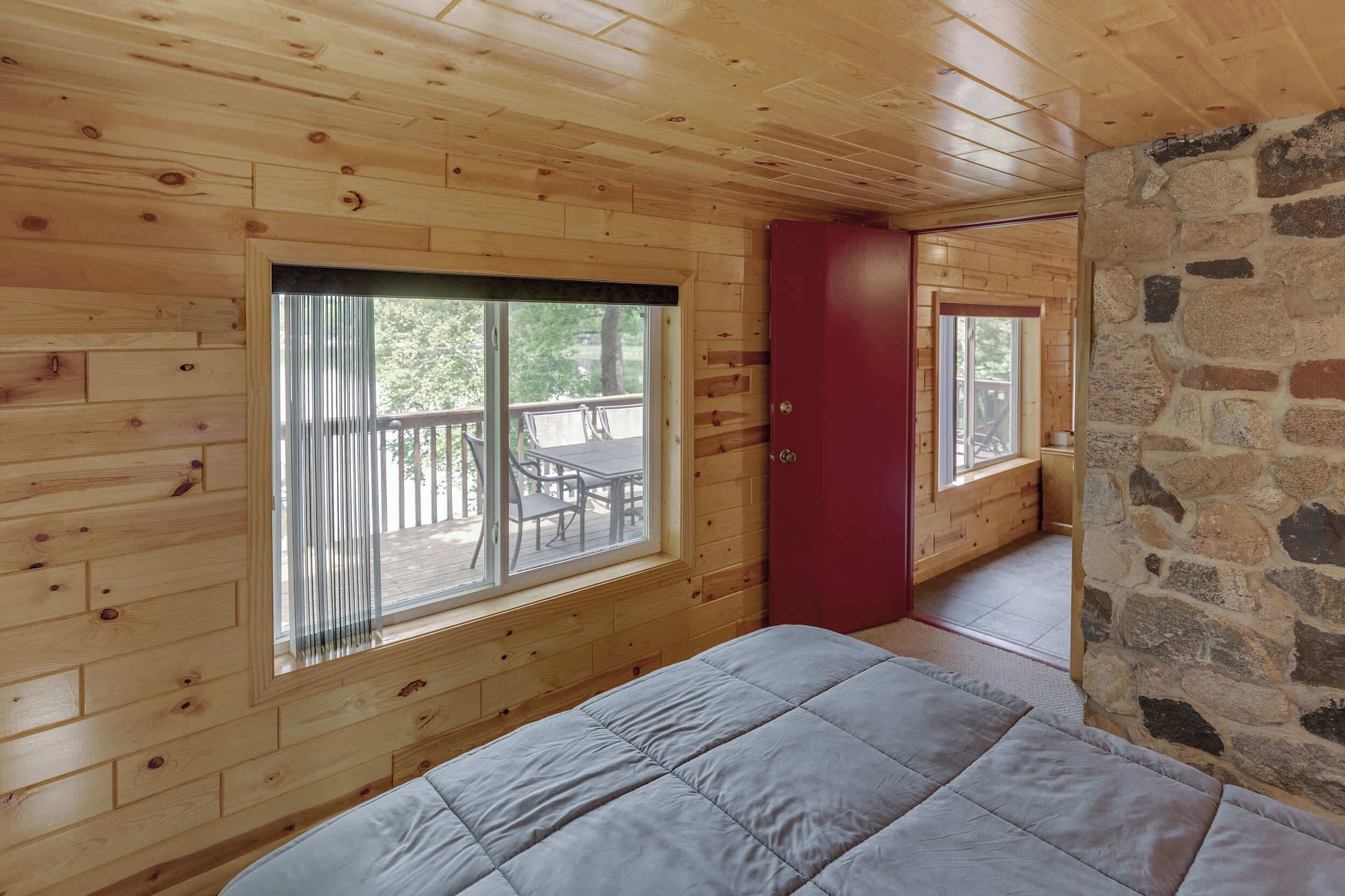 A wooden cabin room with a bed, a red door, and large windows overlooking a deck.
