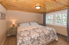 A bedroom with a wooden ceiling, a large bed with a floral bedspread, a nightstand with a lamp, and a window with curtains.