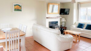 Ett vardagsrum med en vit soffa, ett matbord i trä med stolar, en öppen spis, en tv som är monterad på väggen och ett fönster med gardiner.