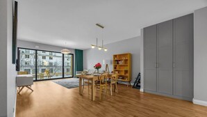 A spacious living room with a wooden floor, a dining area with a table and chairs, a bookshelf, and a large sliding door closet.