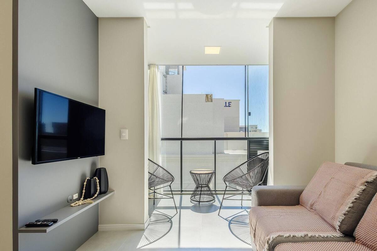 Een moderne woonkamer met een flatscreen televisie, een bank, een glazen tafel met stoelen, en een balkon met uitzicht op gebouwen.