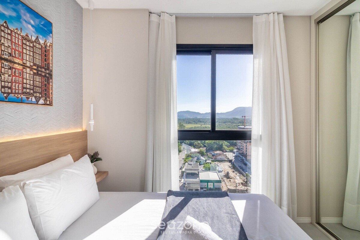 A hotel room with a bed, a painting of buildings, a window with a view of a town, and a nightstand with a lamp.