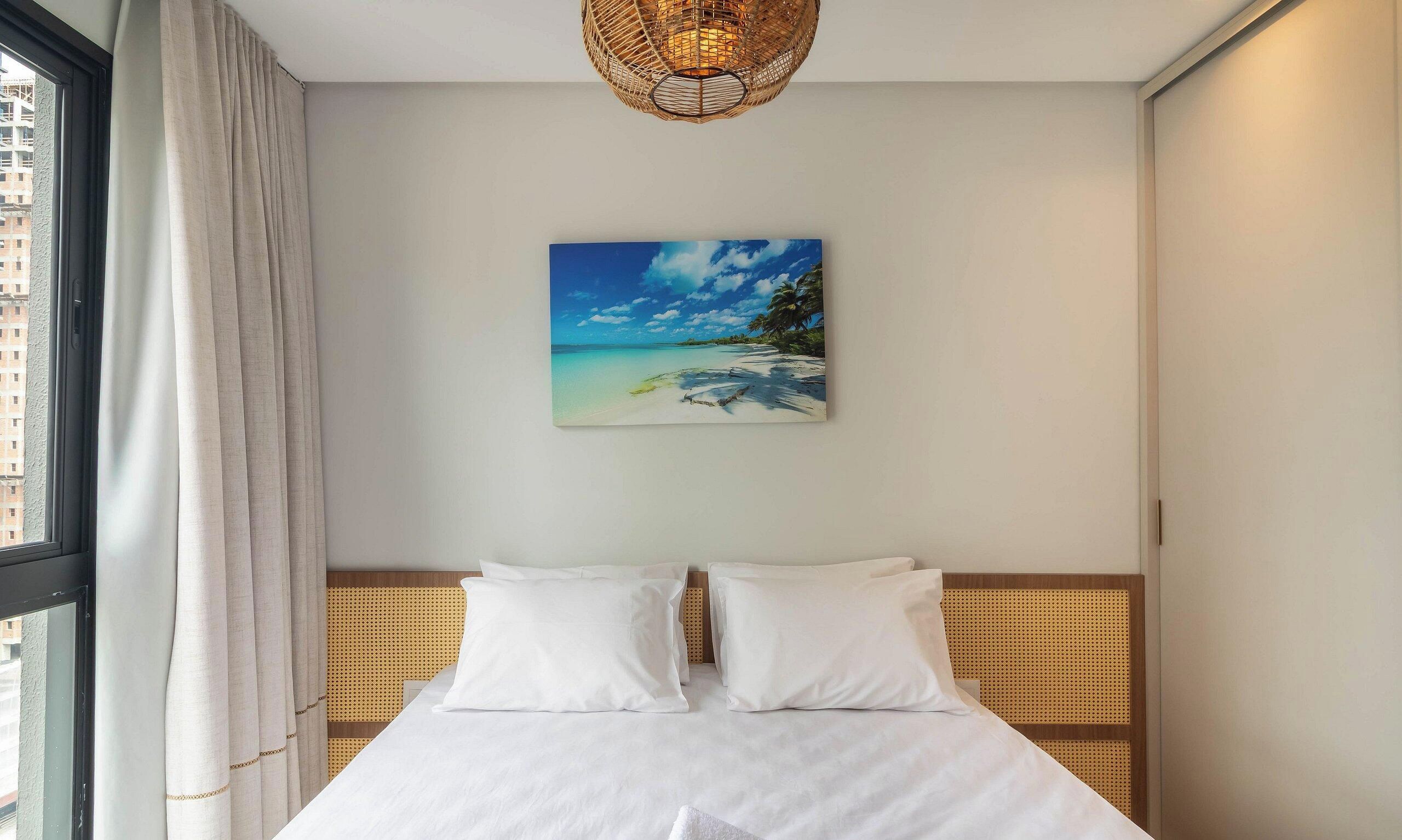 A hotel room with a bed, a painting of a beach, and a window with curtains.