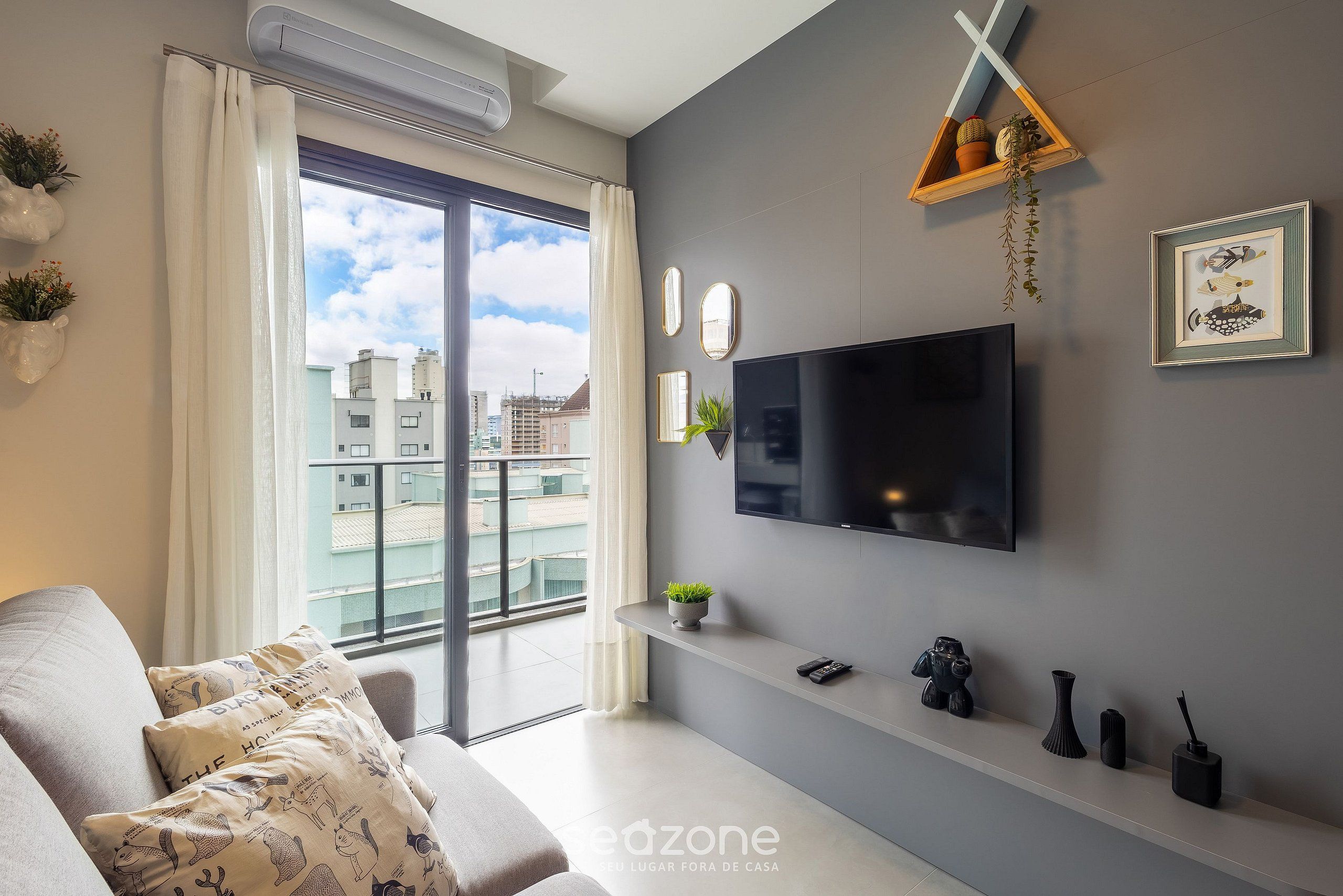 A modern living room with a large TV, a sofa with patterned cushions, a balcony view, and a mounted shelf with decorative items.