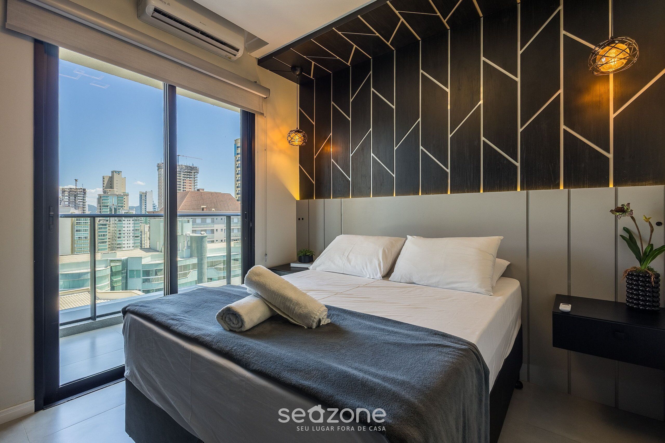Quarto moderno com uma janela grande, uma cama com toalha enrolada, mesa de cabeceira e planta decorativa.