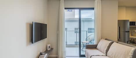 A modern living room with a flat-screen TV, a sofa, a small table, and a balcony with a view of a building.