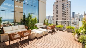 Un área lounge moderna al aire libre con terraza de madera, sofás blancos, sillones de mimbre y plantas en macetas.