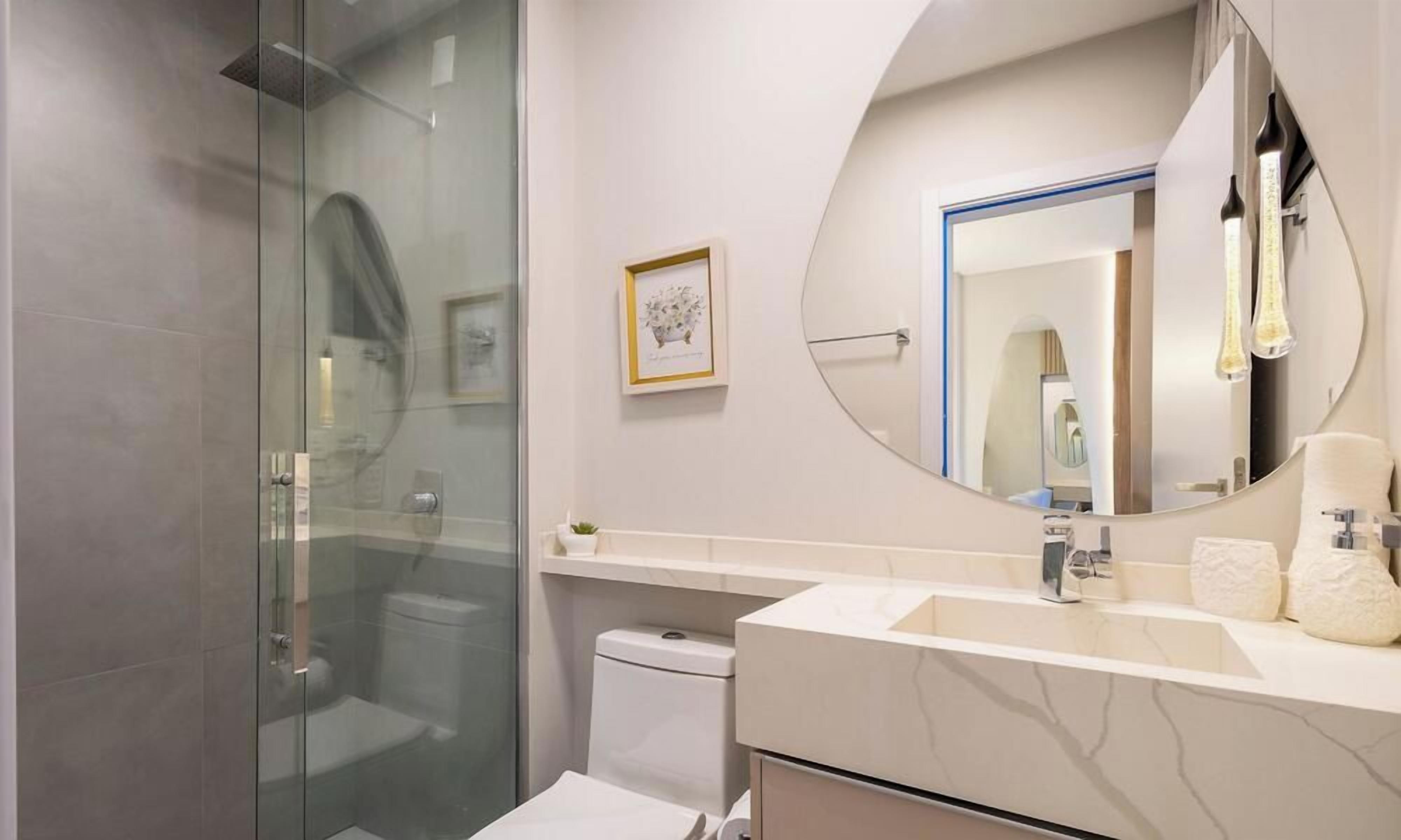 A modern bathroom with a glass shower enclosure, a round mirror, a white sink with a marble countertop, and a small framed picture on the wall.