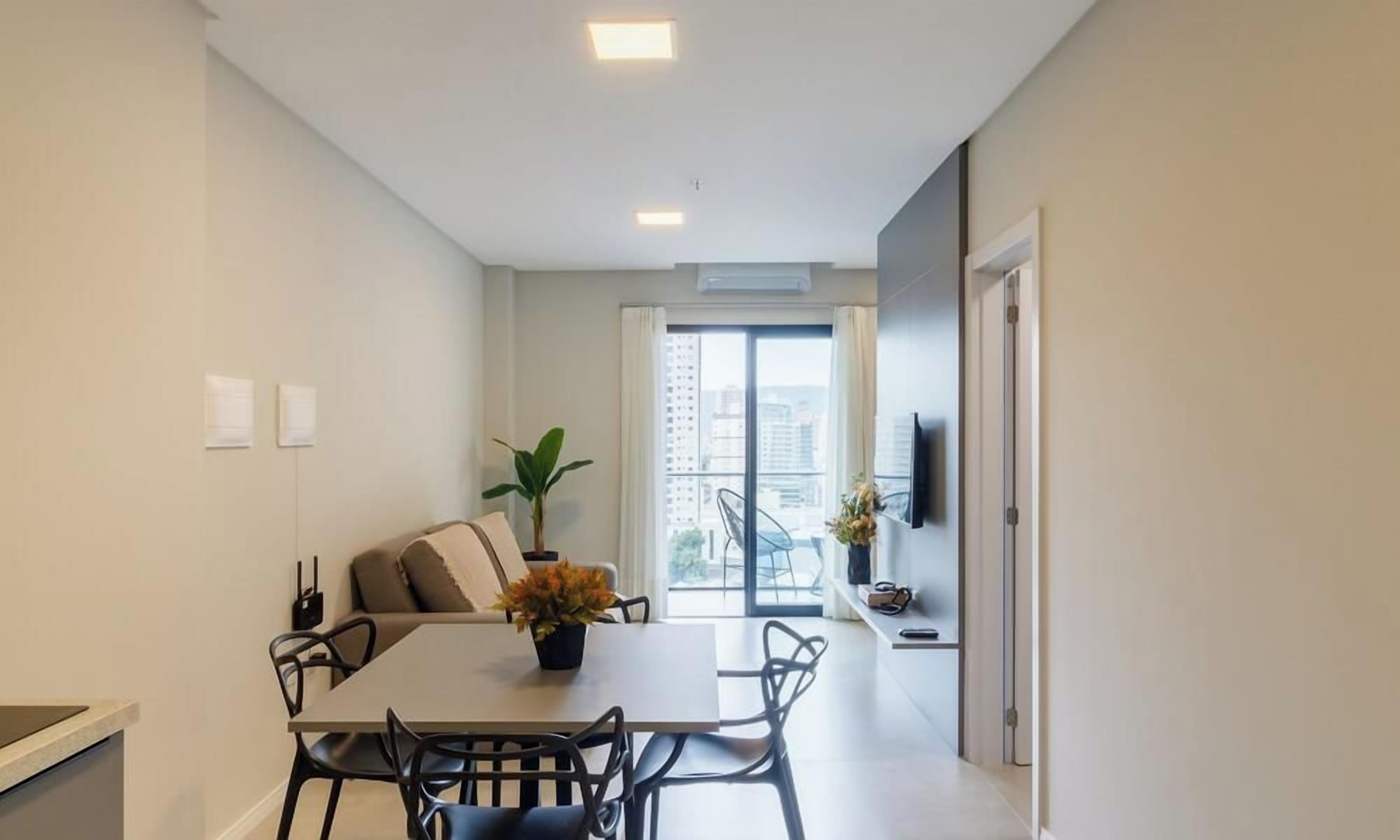 A modern apartment with a dining area, a sofa, a potted plant, and a view of the city through a large window.