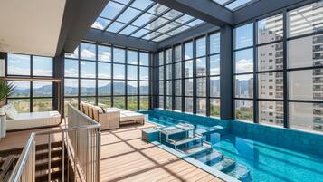 A rooftop infinity pool with a glass ceiling, wooden deck, and a seating area with a view of the cityscape.