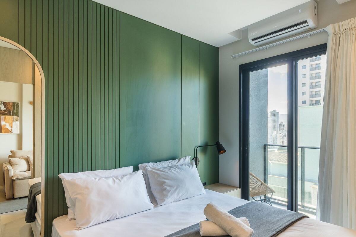 Une chambre d’hôtel comprenant un lit, une table de chevet, une lampe, une fenêtre avec des rideaux et un appareil de climatisation.