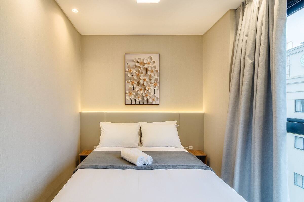 Een hotelkamer met een bed, een nachtkastje, een wandlamp en een raam met gordijnen.