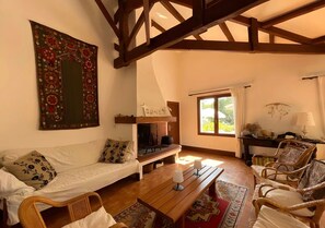 A cozy living room with a wooden ceiling, a sofa with patterned cushions, a wooden table, and a large window with curtains. - Family Home on the Edge of the Forest with Terrace, Private Garden, and Balcony (Le Grand-Village-Plage)