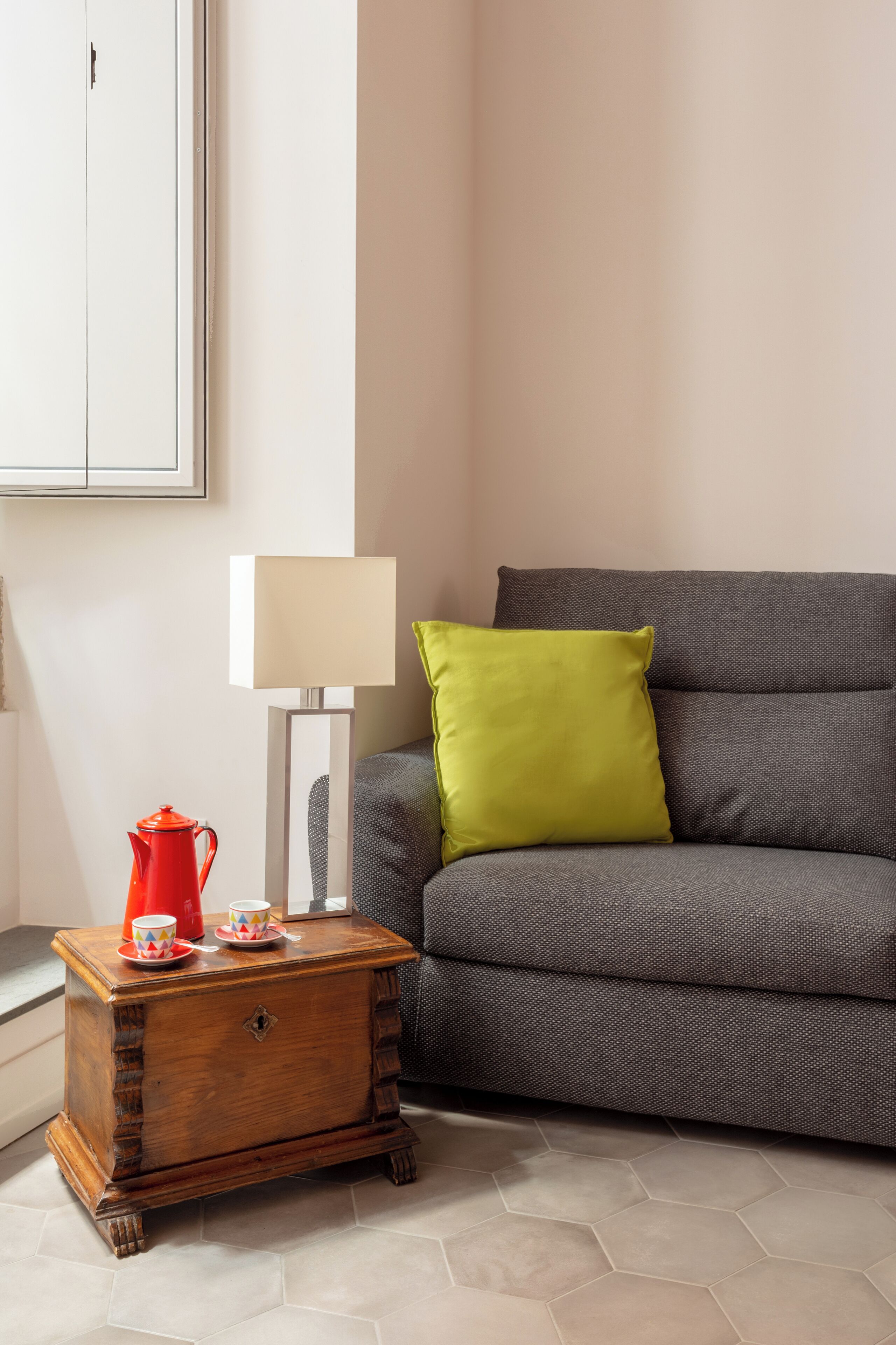 Una sala de estar con un sofá gris, una mesita de madera con una tetera roja y una lámpara blanca.