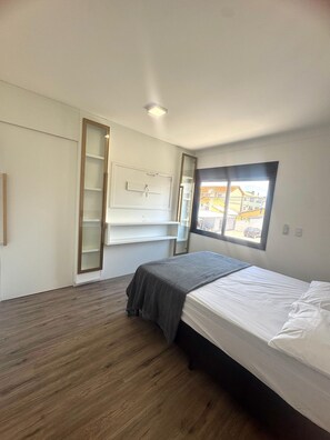 A bedroom with a bed, a window with a view, a shelf unit, and a wooden floor. - Sobrado 00 (Florianópolis)