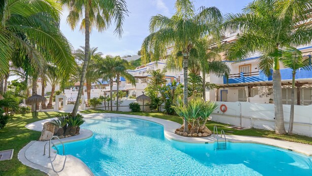 A swimming pool surrounded by palm trees and a building with a blue roof. - Oasis Tropical Apartment with Parking, Pool, and Air Conditioning (Almuñécar)