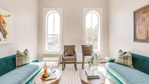 Un salón espacioso con dos sofás verdes, una mesa de centro de madera y dos sillones. Amplias ventanas ofrecen luz natural y vistas al exterior.