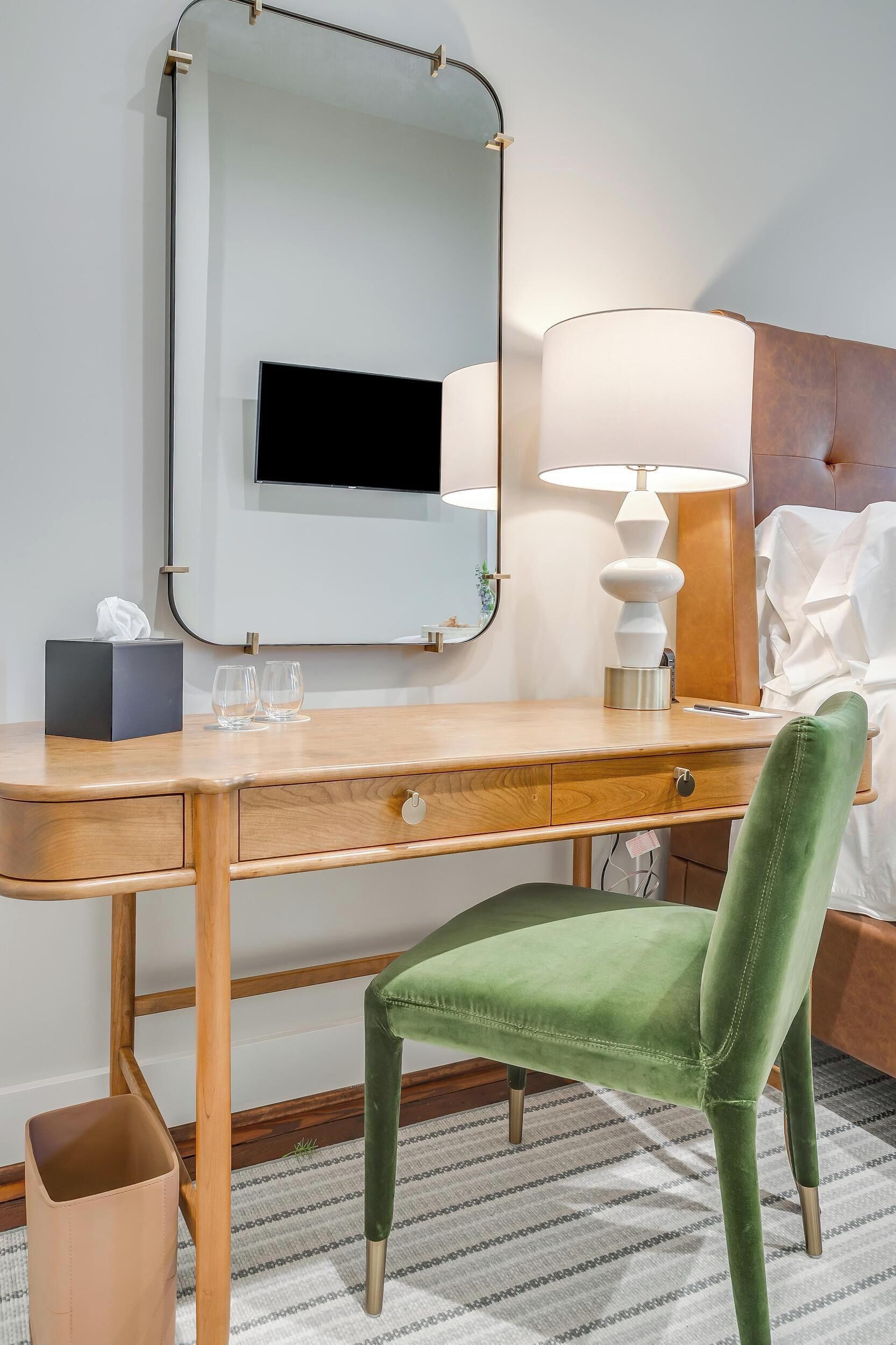 Habitación de hotel con un escritorio de madera, una silla tapizada de verde, una lámpara de noche, un televisor de pantalla plana y una cama con sábanas blancas.