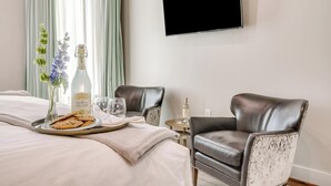 Ein Hotelzimmer mit einem Bett, einem Tablett mit Keksen, einer Flasche Sprudelzitrone und einer Wasserflasche auf einem kleinen Tisch. Es befinden sich zwei Ledersessel und ein an der Wand befestigtes Fernsehgerät dort.