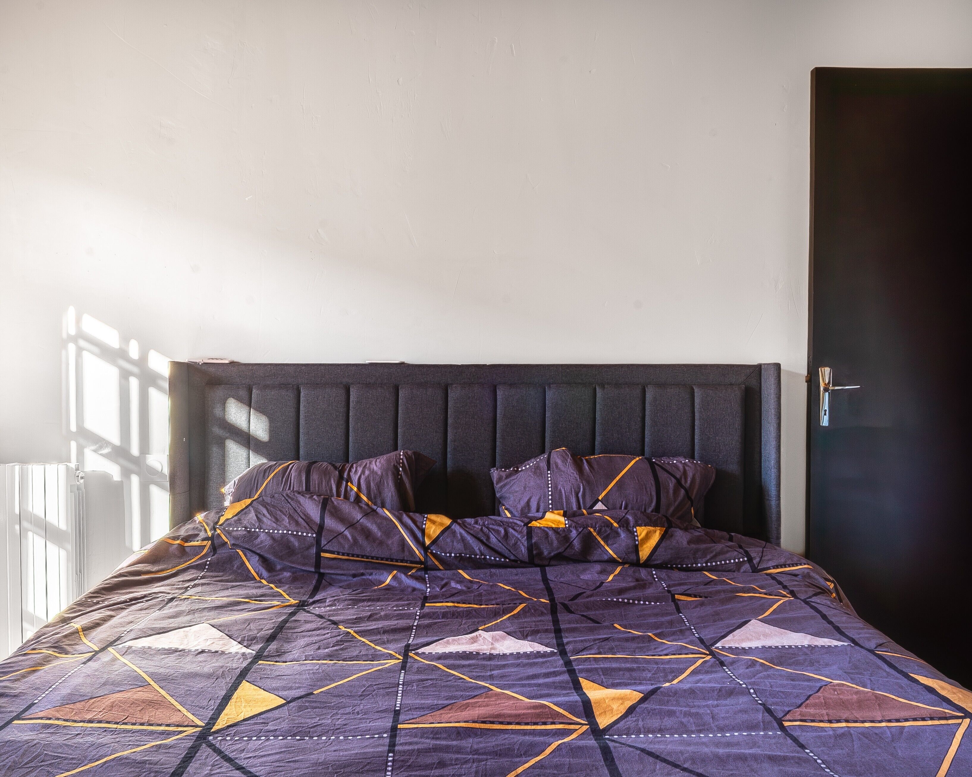 Een bed met een bedspread met geometrisch patroon, een donker hoofdbord en een deur aan de rechterkant.