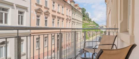 A balcony with two chairs and a table, overlooking a street with buildings and a hill in the background.