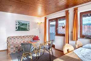 Ein gemütliches Zimmer mit Holzbalkendecke, einem gemusterten Sofa, einem Esstisch mit Stühlen und Blick durch ein Fenster mit Vorhängen.