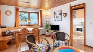 Ett mysigt vardagsrum med ett träpanelat tak, en matplats med bord och stolar, en tv och ett kök som syns genom en öppen dörr.