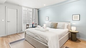 Een hotelkamer met een bed, nachtkastjes, een raam met gordijnen, een deur en een klein tafeltje met een vaas.