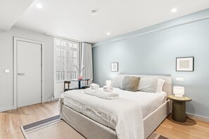 Ein Hotelzimmer mit Bett, Nachttischen, einem Fenster mit Vorhängen, einer Tür und einem kleinen Tisch mit einer Vase.