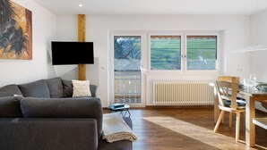 A living room with a sofa, a TV, a dining table with chairs, a window with a view of a green landscape, and a radiator. - Apartment Wiesenblick with Balcony and Wi-Fi (Horben)