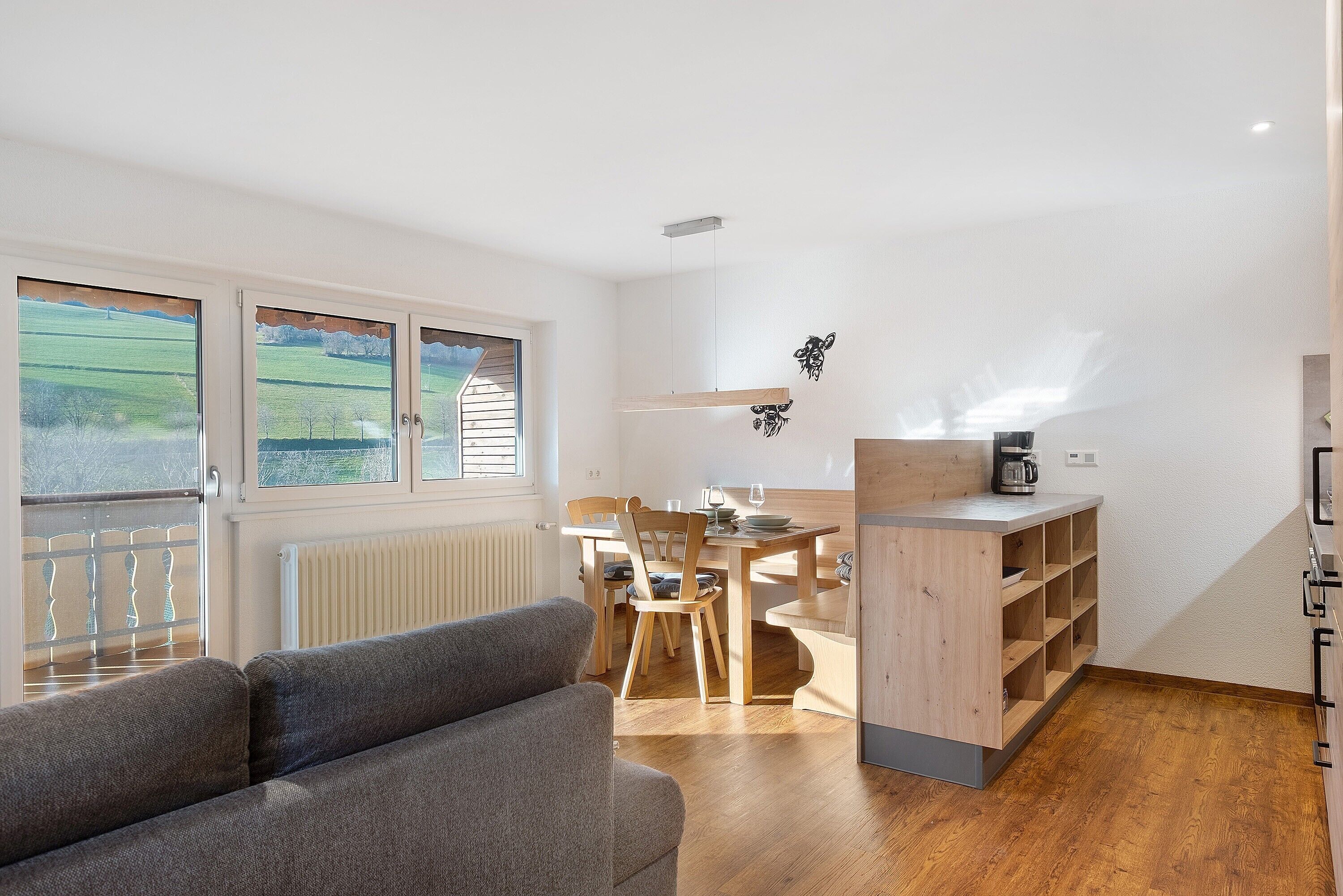A modern room with a wooden dining table, chairs, a sofa, and a kitchen area with a coffee maker.