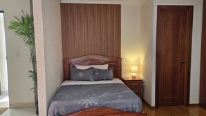 Een slaapkamer met een houten bed, een nachtkastje met een lamp, een houten deur en een potplant.