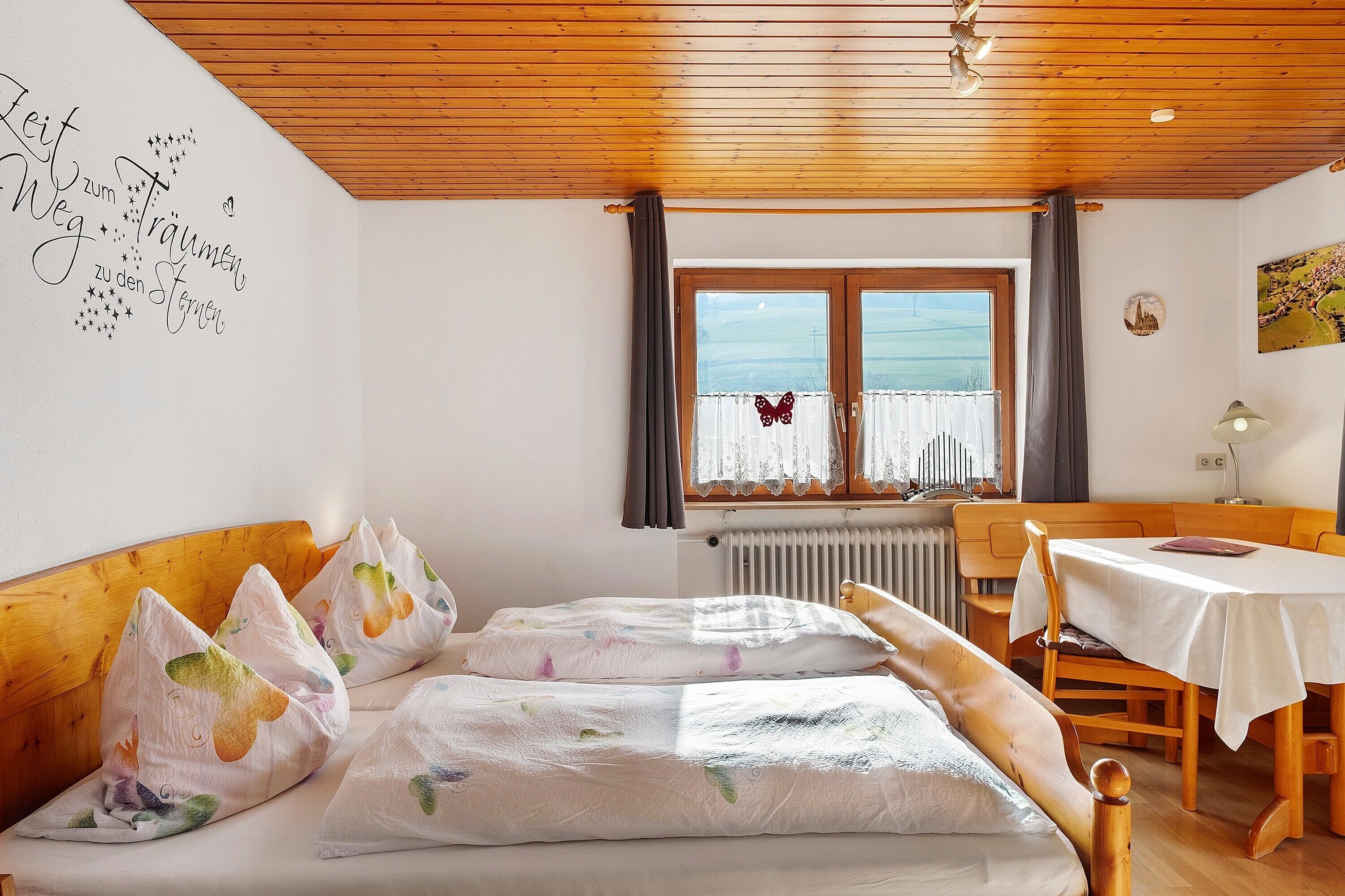 Una habitación con techo de madera, una cama con almohadas coloridas, una mesa con mantel blanco y una ventana con cortinas.