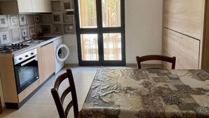 Cocina compacta con estufa, horno y lavadora, una mesa de comedor con sillas y un ventanal con cortinas.