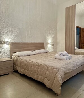 Ein ordentlich bezogenes Bett mit einer beigen Decke, ein Nachttisch mit Schubladen, eine Wandleuchte und ein Spiegel, der eine Holztür reflektiert.