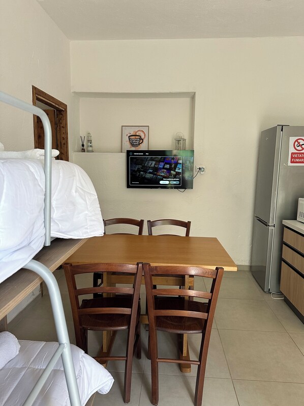 Een kleine kitchenette met een koelkast, een aan de muur bevestigde televisie, een houten tafel met stoelen en een stapelbed met wit beddengoed.