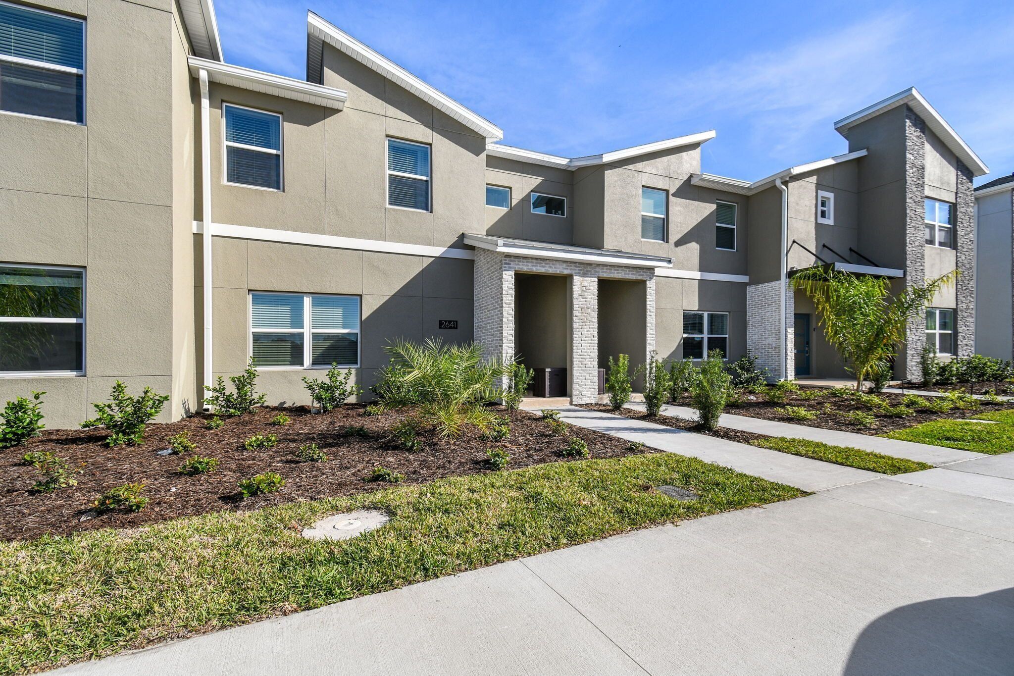 A modern multi-story residential building with a neutral color palette, a landscaped front yard, and a clear sky.