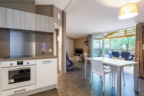 Moderni keittiö, jossa on integroidut kodinkoneet, ruokailutila pöytineen ja tuoleineen sekä suuri ikkuna, josta avautuu näkymä vehreään maisemaan.