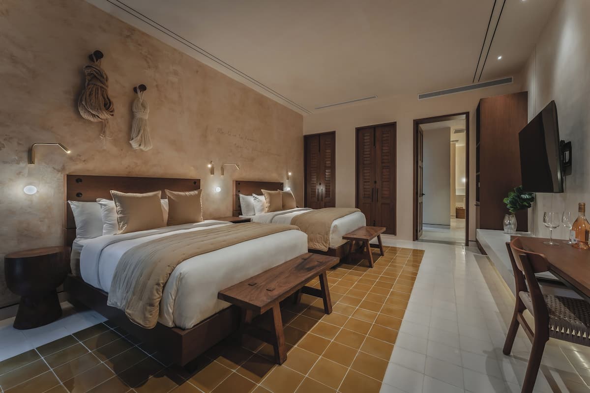 A hotel room with two beds, a wooden bench, a dining table, and a TV.